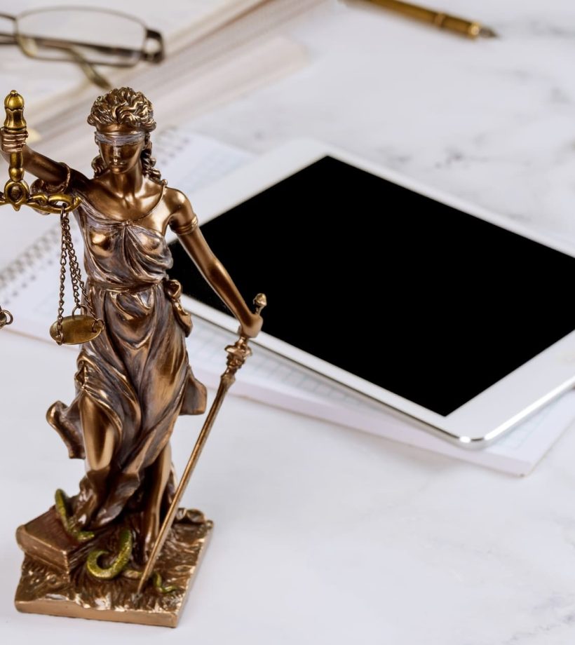 Estatua de la justicia sobre mesa de mármol. Atrás se ven una tablet, unas gafas sobre unos informes, y unas esposas.